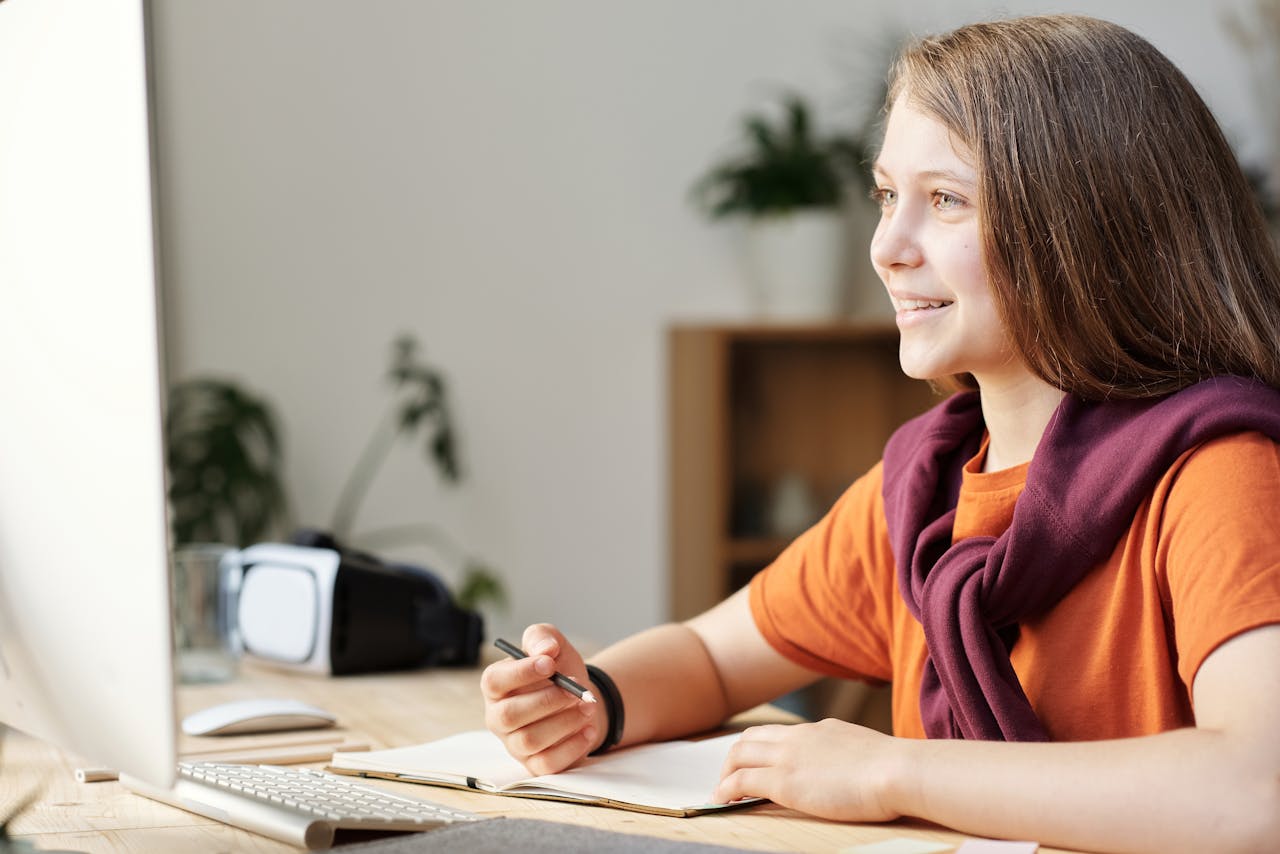 Services A smiling teenage girl engages in online learning at her home workspace, using modern technology.